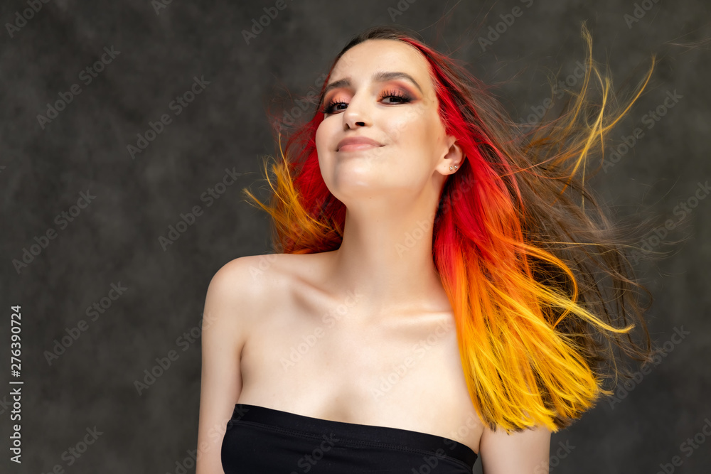 A close-up portrait photo of a fashionable hairstyle red-yellow in studio on a gray background. The pretty brunette model with beautiful make-up has beautiful flowing colorful hair.