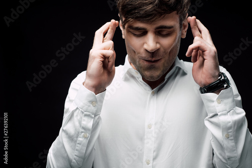 businessman with closed eyes holding crossed fingers isolated on black, human emotion and expression concept photo