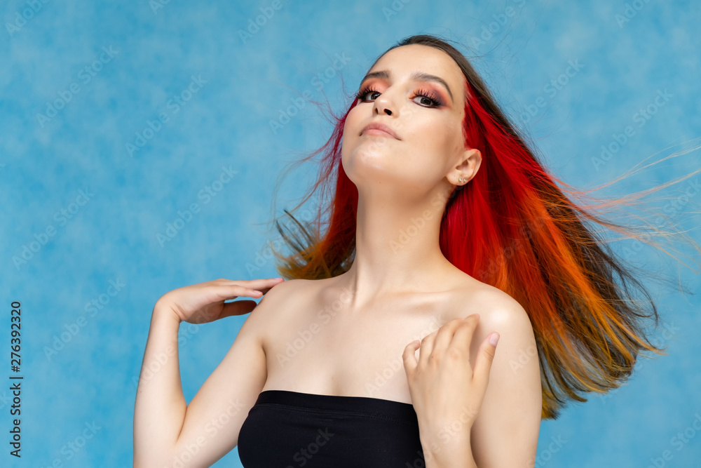 A close-up portrait photo of a fashionable hairstyle red-yellow in studio on a blue background. The pretty brunette model with beautiful make-up has beautiful flowing colorful hair.