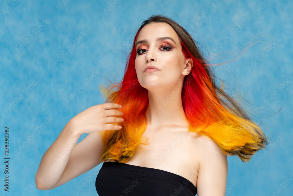 A close-up portrait photo of a fashionable hairstyle red-yellow in studio on a blue background. The pretty brunette model with beautiful make-up has beautiful flowing colorful hair. A close-up portrait photo of a fashionable hairstyle red-yellow in studio on a blue background. The pretty brunette model with beautiful make-up has beautiful flowing colorful hair.