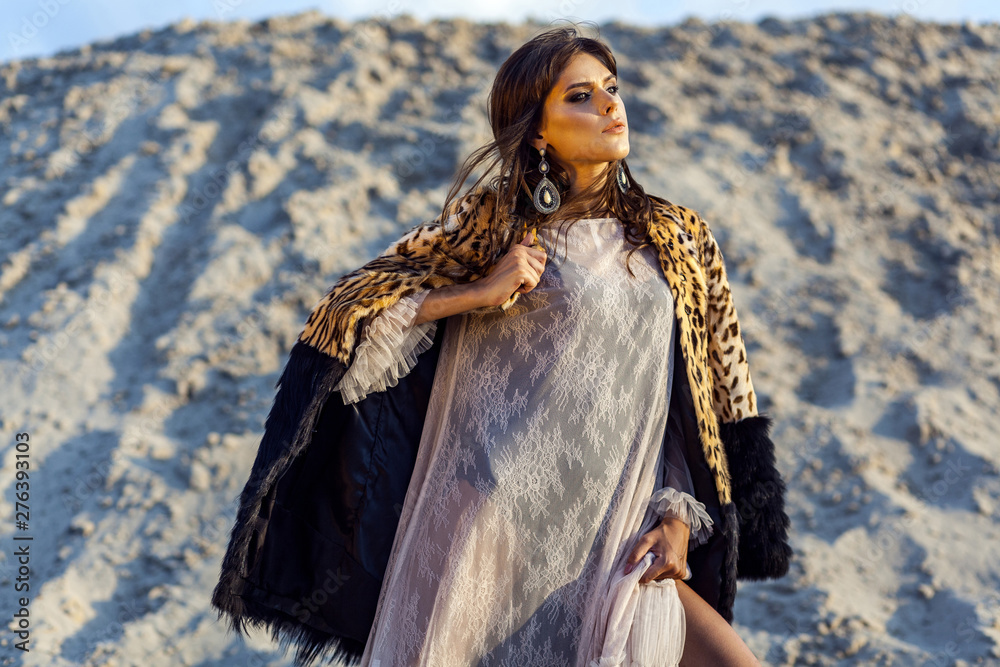 Attractive brunette woman in translucent beach cover up with leopard fur coat posing on sandy beach at sunset. holding coat and looking away with passion. Fashion outdoor shot in the summertime.
