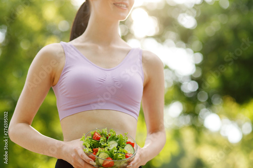 Beautiful slim brunette eating salad over green natural background. Female fitness model at the park. Woman hands holding fresh summer salad with raw vegetables cucumbers tomatoes lettuce in bowl.