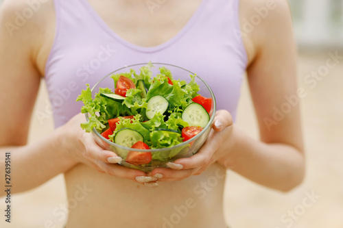 Beautiful slim girl eating salad. Female fitness model on diet. Woman hands holding fresh summer salad with raw vegetables cucumbers tomatoes lettuce in bowl.