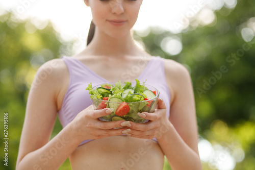 Beautiful slim brunette eating salad over green natural background. Female fitness model at the park. Woman hands holding fresh summer salad with raw vegetables cucumbers tomatoes lettuce in bowl.