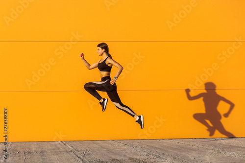 Young satisfied sporty beautiful woman with fit body jumping and running, hurry up against orange wall background. Gymnast jumping high with toothy smile, full length, outdoor