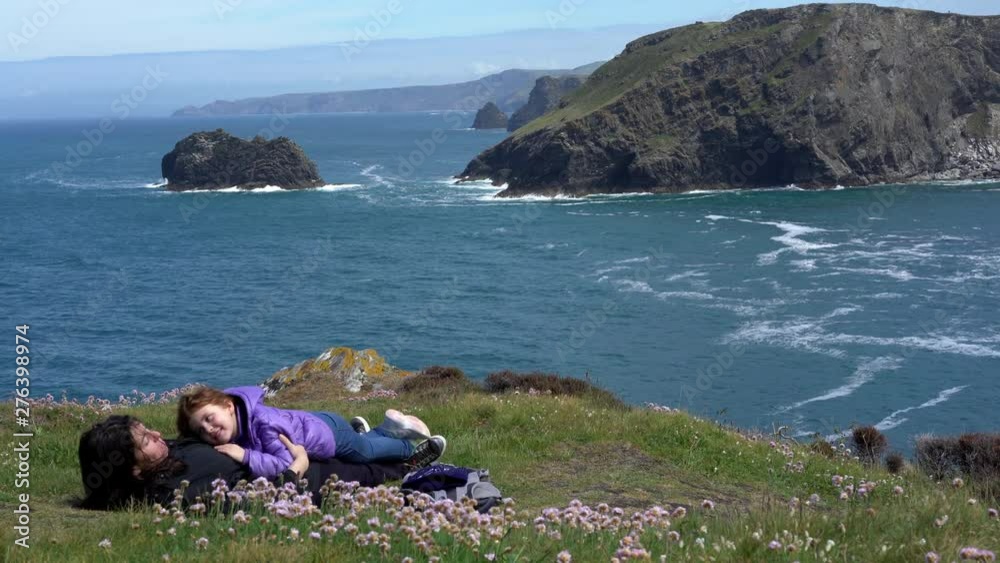 Happy mother and little daughter lying on the grass on top of a cliff overlooking the ocean in Cornwall on a beautiful spring sunny day. 4k footage