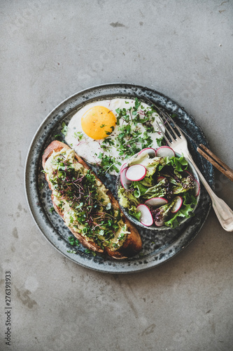 Healthy trendy breakfast. Flat-lay of avocado toast on sourdough bread with herbs and sprouts, fried egg with coriander and salad in plate over grey concrete background, top view. Clean eating concept