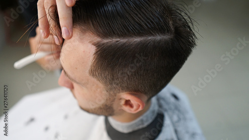 Interior shot of working process in modern barbershop. Close-up portrait of attractive young man getting trendy haircut. Male hairdresser serving client, making haircut using metal scissors and comb