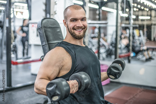 Portrait of sporty healthy strong muscle charismatic happy smiling handsome man bodybuilder hard training workout in well-equipped gym doing biceps exercises with dumbbells in black t-shirt outfit