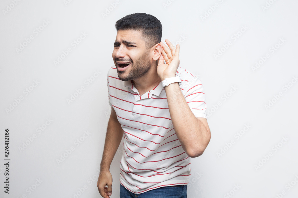 What? I can not hear you. Portrait of attentive handsome bearded young man in striped t-shirt standing with hand on ear and want to hear something. indoor studio shot, isolated on white background. What? I can not hear you. Portrait of attentive handsome bearded young man in striped t-shirt standing with hand on ear and want to hear something. indoor studio shot, isolated on white background.