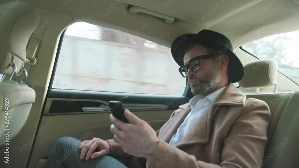 Close-up of handsome fashion man in retro outfit using a smartphone in the car. Serious thoughtful businessman driving a car through city on a sunny autumn day.