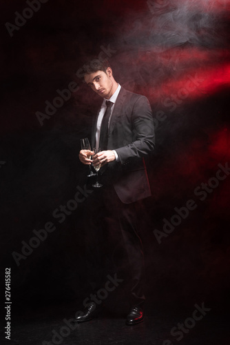 full length view of handsome man in formal wear holding wineglasses in smoke on black
