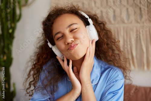 Funny curly young woman siting with closed eyes, listening favorite song in headphones, enjoying the music and Sundays morning.