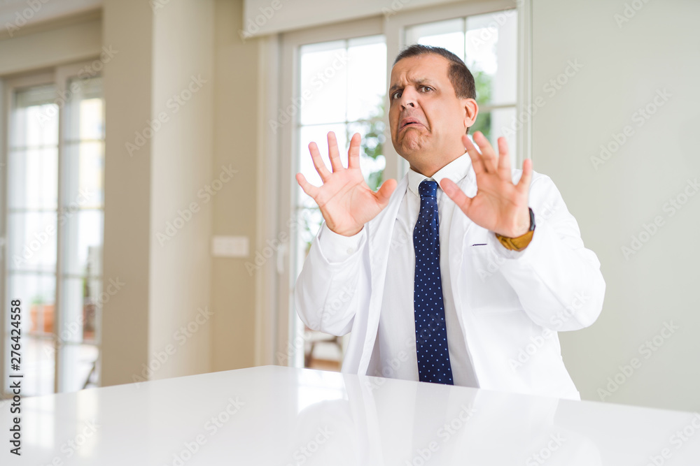 Middle age doctor man wearing medical coat at the clinic afraid and terrified with fear expression stop gesture with hands, shouting in shock. Panic concept.
