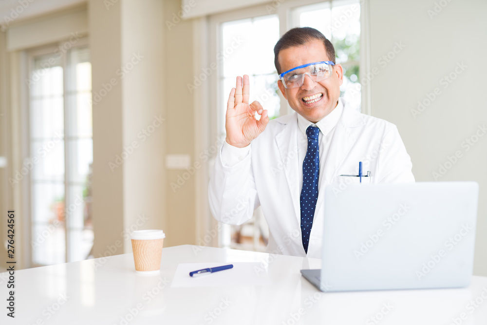 Middle age scientist man wearing professional glasses working using laptop doing ok sign with fingers, excellent symbol