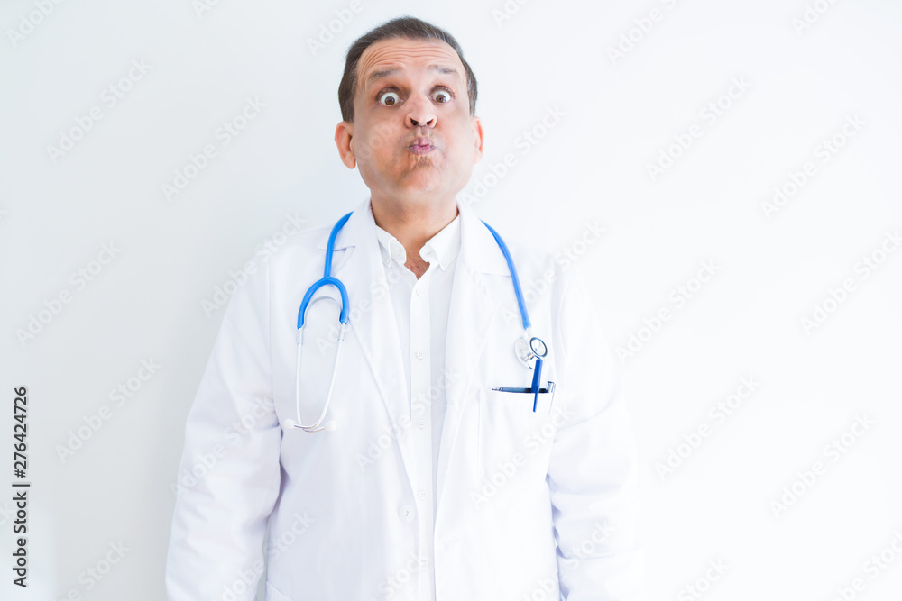 Middle age doctor man wearing stethoscope and medical coat over white background puffing cheeks with funny face. Mouth inflated with air, crazy expression.