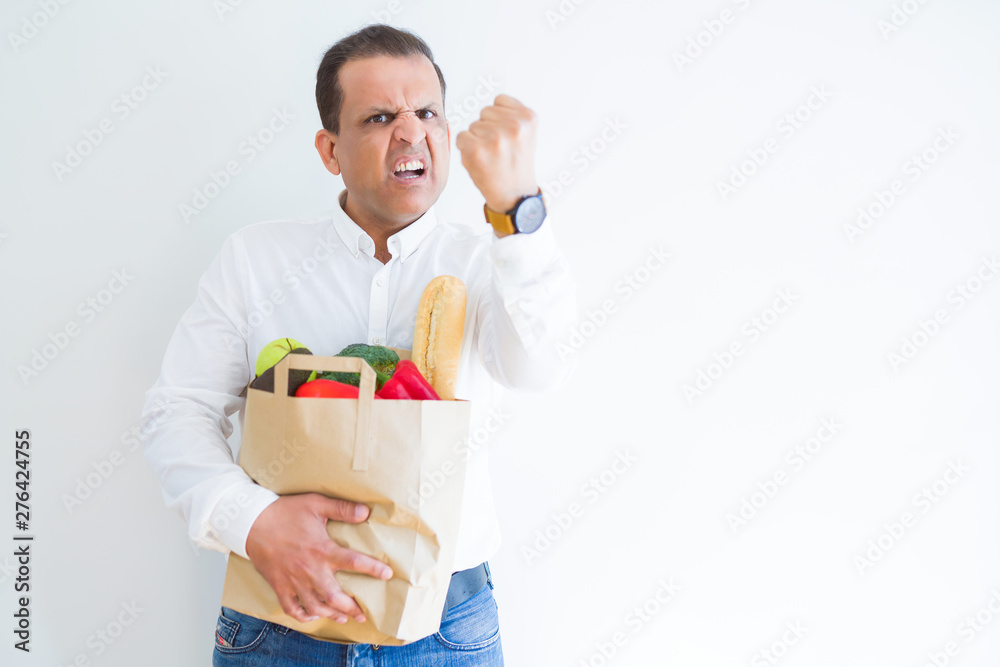 Middle age man holding groceries shopping bag over white background annoyed and frustrated shouting with anger, crazy and yelling with raised hand, anger concept