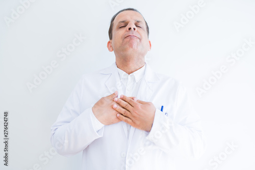 Middle age doctor man wearing medical coat over white background smiling with hands on chest with closed eyes and grateful gesture on face. Health concept.