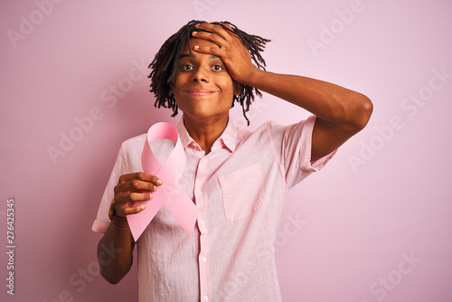 Afro american man with dreadlocks holding cancer ribbon over isolated pink background stressed with hand on head  shocked with shame and surprise face  angry and frustrated. Fear and upset 