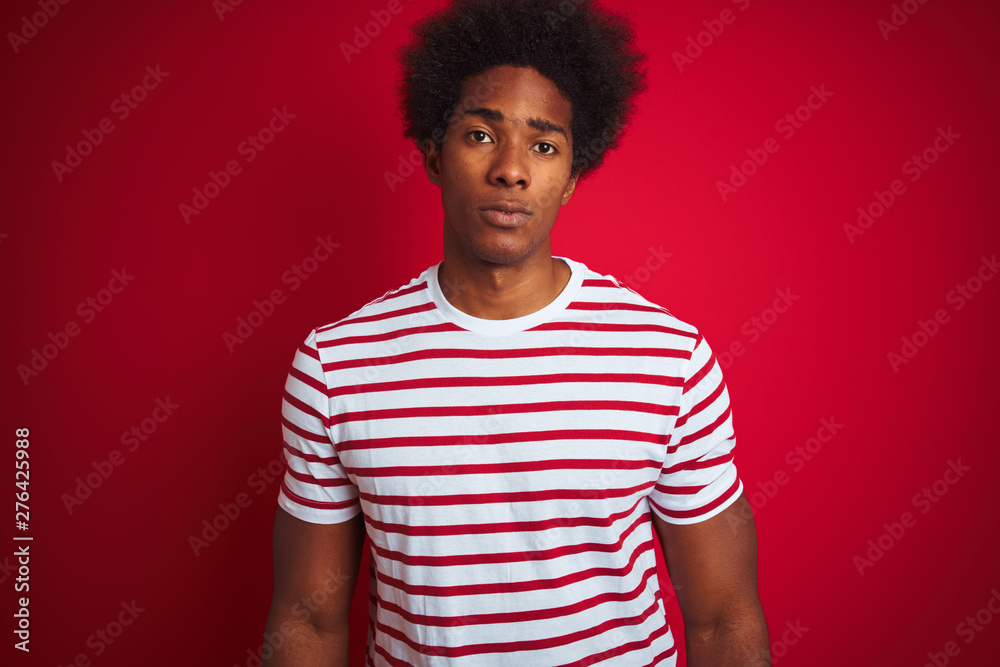Young african american man with afro hair wearing striped t-shirt over isolated red background Relaxed with serious expression on face. Simple and natural looking at the camera. Young african american man with afro hair wearing striped t-shirt over isolated red background Relaxed with serious expression on face. Simple and natural looking at the camera.