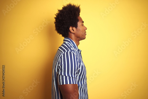 American man with afro hair wearing striped shirt standing over isolated yellow background looking to side, relax profile pose with natural face with confident smile.