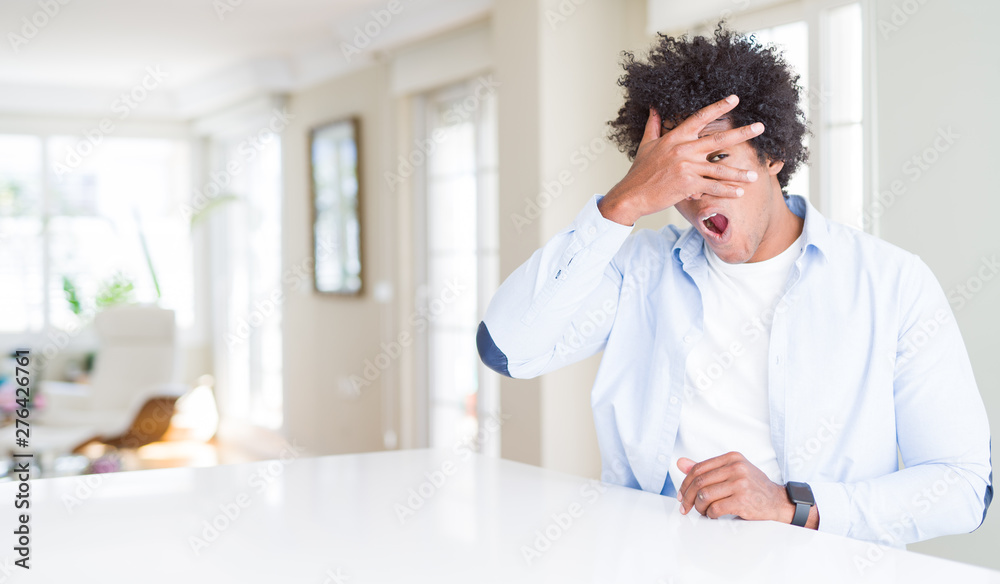 African American man at home peeking in shock covering face and eyes with hand, looking through fingers with embarrassed expression.