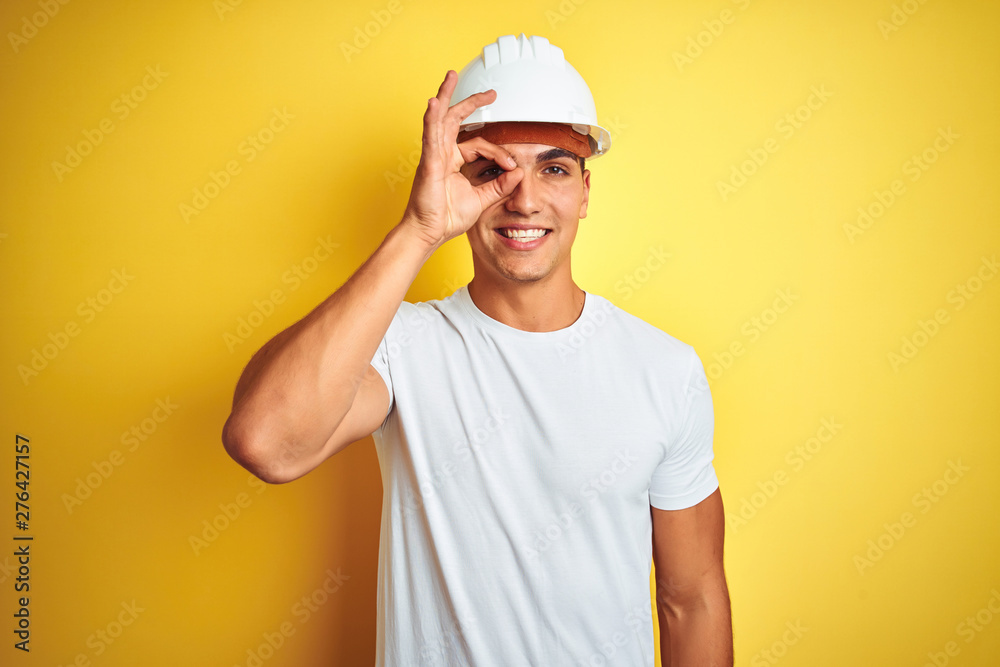 Young handsome man wearing construction helmet over yellow isolated background doing ok gesture with hand smiling, eye looking through fingers with happy face.
