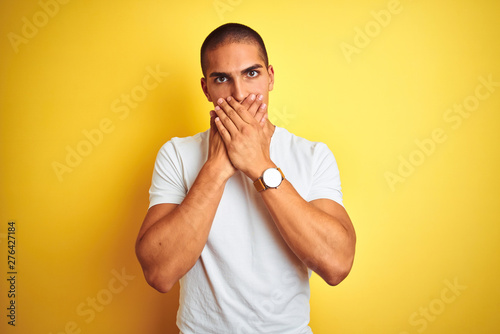 Young caucasian man wearing casual white t-shirt over yellow isolated background shocked covering mouth with hands for mistake. Secret concept.