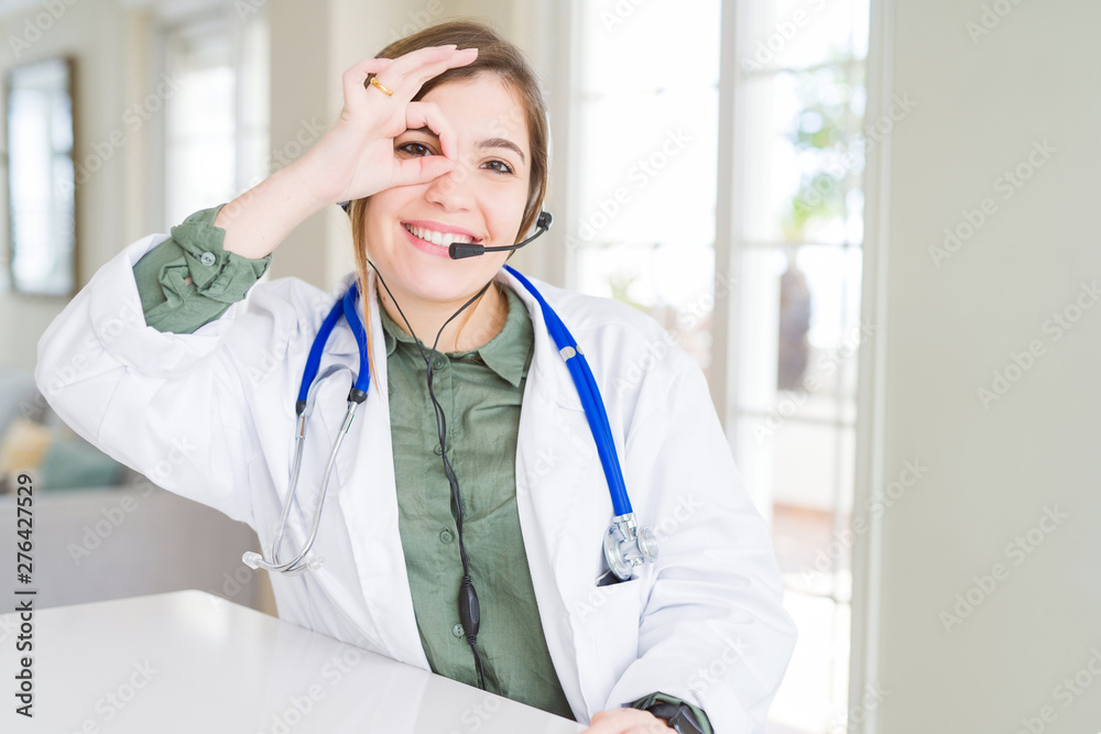 Beautiful young doctor woman wearing headset doing ok gesture with hand smiling, eye looking through fingers with happy face. Beautiful young doctor woman wearing headset doing ok gesture with hand smiling, eye looking through fingers with happy face.