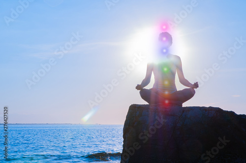 Woman is meditating with glowing seven chakras on stone at sunset. Silhouette of woman is practicing yoga on the beach.