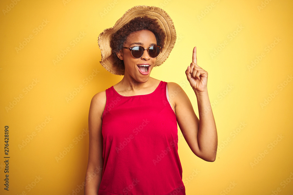 African american woman wearing summer hat and sunglasses over yellow isolated background pointing finger up with successful idea. Exited and happy. Number one. African american woman wearing summer hat and sunglasses over yellow isolated background pointing finger up with successful idea. Exited and happy. Number one.