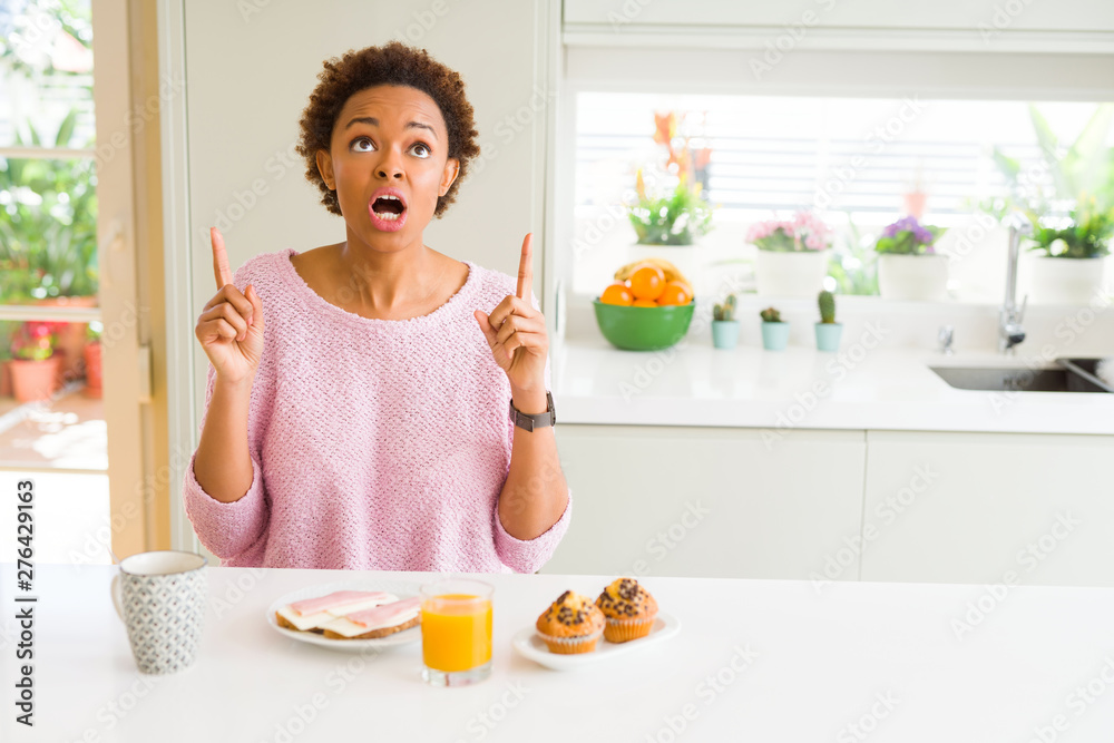 Young african american woman eating breaksfast in the morning at home amazed and surprised looking up and pointing with fingers and raised arms.