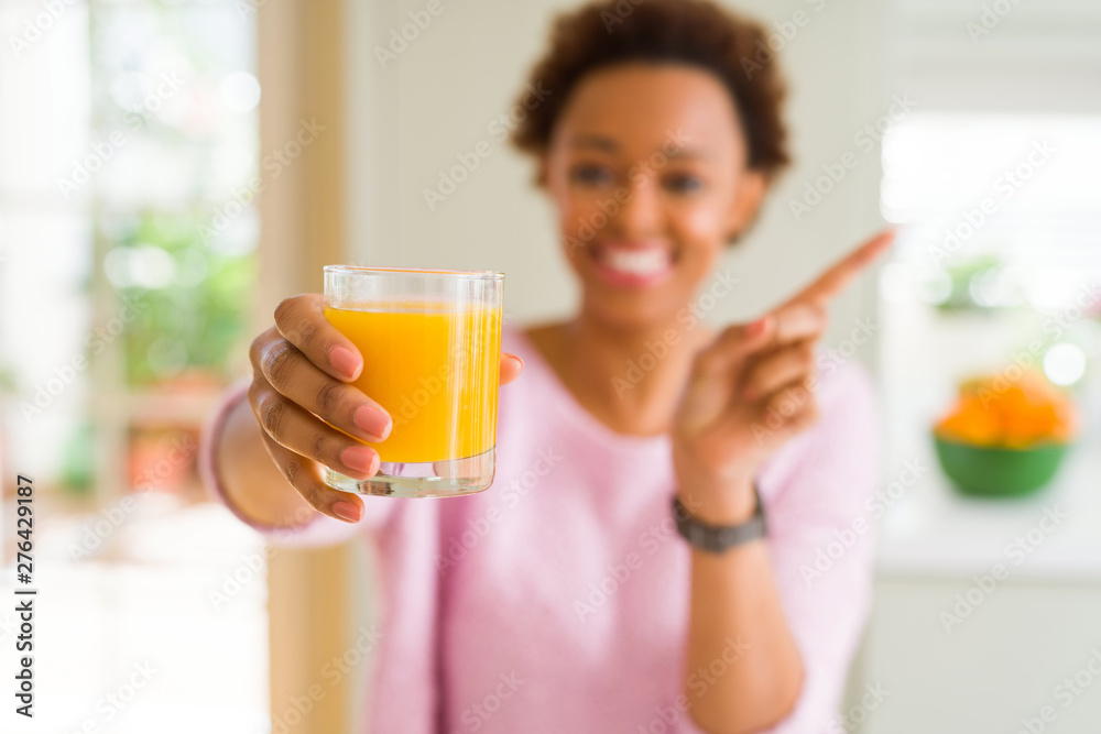 Young african american woman driking orange juice at home very happy pointing with hand and finger to the side Young african american woman driking orange juice at home very happy pointing with hand and finger to the side
