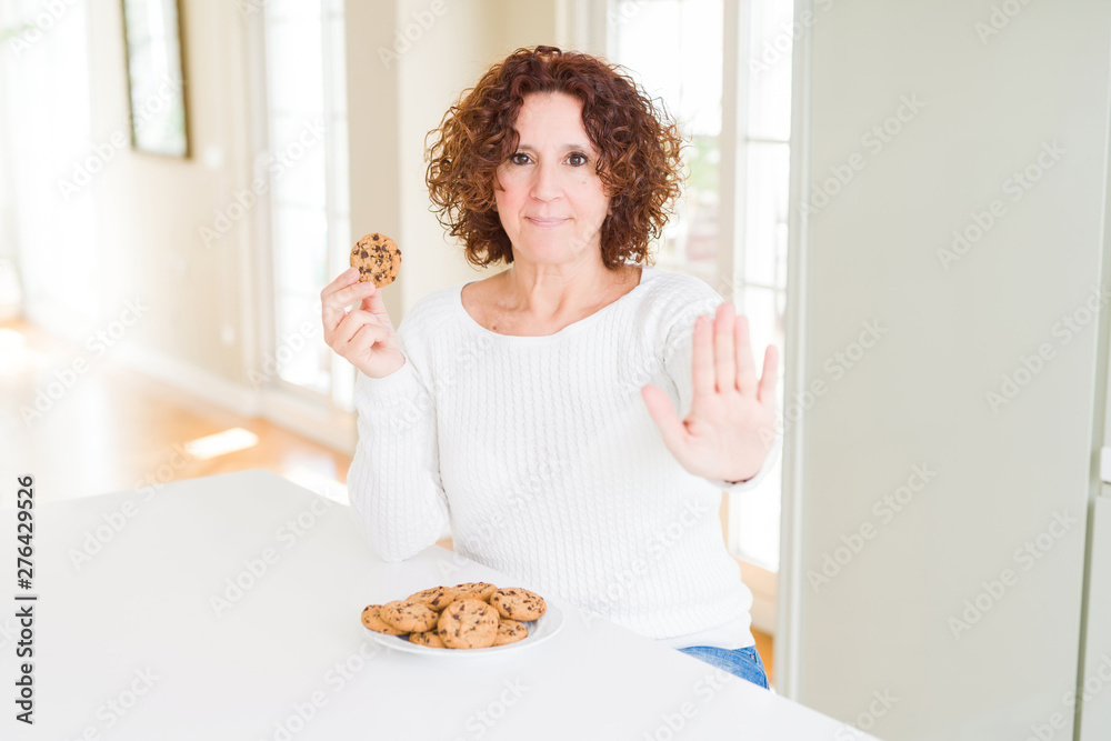 Senior woman eating chocolate chips cookies at home with open hand doing stop sign with serious and confident expression, defense gesture Senior woman eating chocolate chips cookies at home with open hand doing stop sign with serious and confident expression, defense gesture