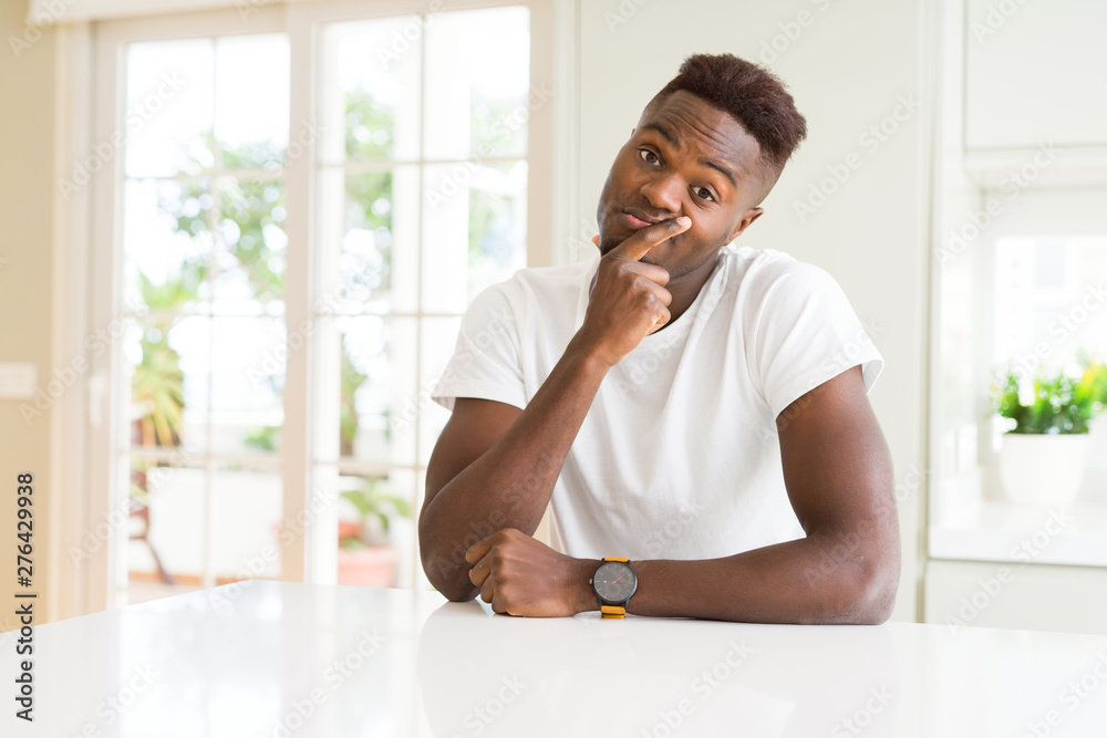 Handsome african american man on white table at home looking confident at the camera with smile with crossed arms and hand raised on chin. Thinking positive. Handsome african american man on white table at home looking confident at the camera with smile with crossed arms and hand raised on chin. Thinking positive.