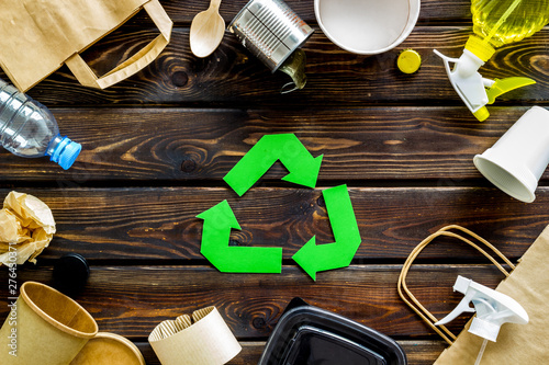 Green recycling sign with waste materials, paper bag and cup, plastic bottles, flatware on wooden background top view