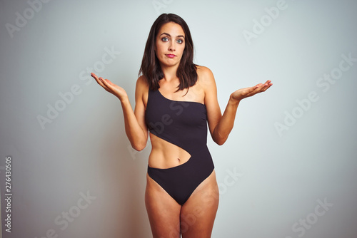 Beautiful woman wearing bikini swimwear over white isolated background clueless and confused expression with arms and hands raised. Doubt concept.