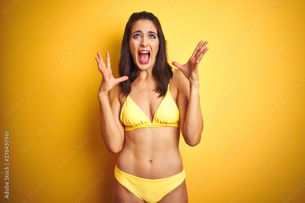 Beautiful woman wearing yellow bikini on summer over isolated yellow background celebrating mad and crazy for success with arms raised and closed eyes screaming excited. Winner concept Beautiful woman wearing yellow bikini on summer over isolated yellow background celebrating mad and crazy for success with arms raised and closed eyes screaming excited. Winner concept