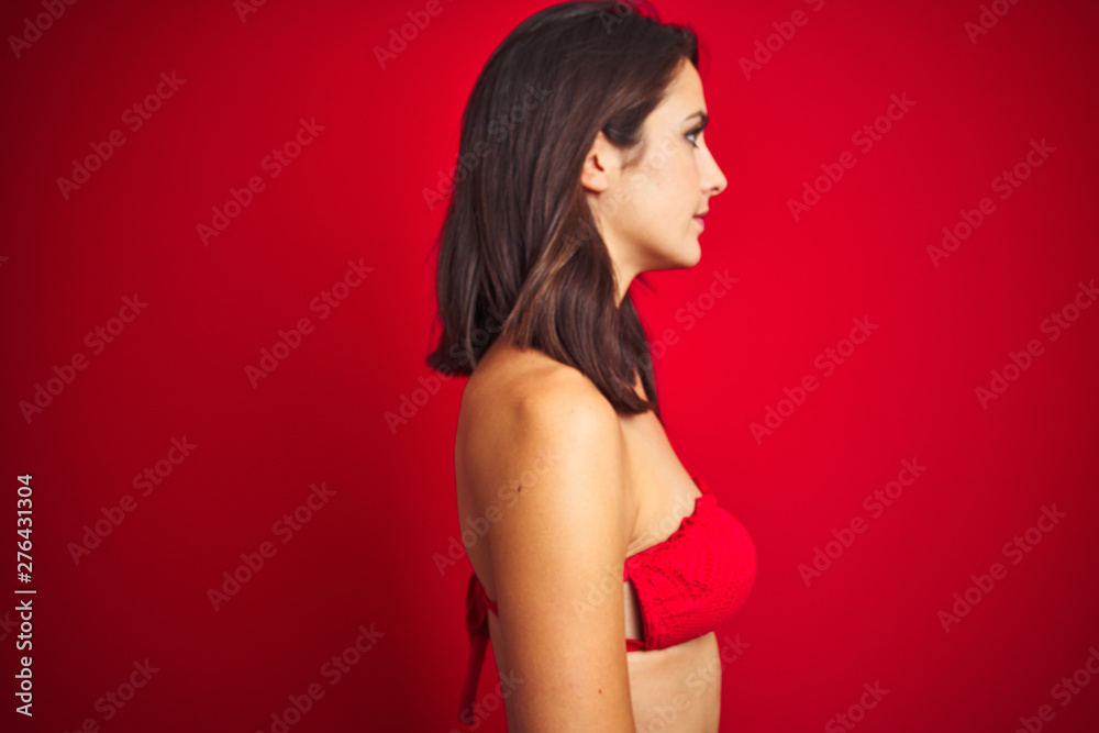 Beautiful woman wearing bikini swimwear over red isolated background looking to side, relax profile pose with natural face with confident smile. Beautiful woman wearing bikini swimwear over red isolated background looking to side, relax profile pose with natural face with confident smile.