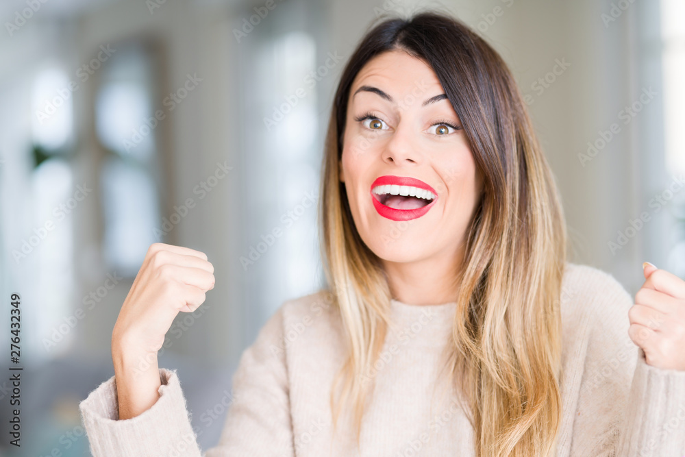 Young beautiful woman wearing winter sweater at home celebrating surprised and amazed for success with arms raised and open eyes. Winner concept. Young beautiful woman wearing winter sweater at home celebrating surprised and amazed for success with arms raised and open eyes. Winner concept.