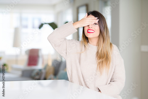 Young beautiful woman wearing winter sweater at home smiling and laughing with hand on face covering eyes for surprise. Blind concept.