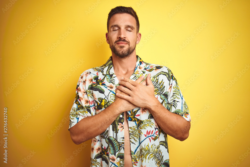 © Krakenimages.com - Handsome man wearing summer hawaiian flowers shirt over yellow isolated background smiling with hands on chest with closed eyes and grateful gesture on face. Health concept.