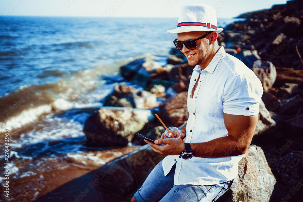 The man at the resort in a white shirt and sunglasses, hat sitting on a rock, drink cocktail and talking on a cell phone on the sea background.
