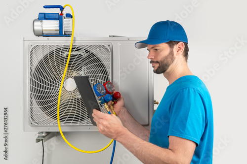Installation service repair of an air conditioner outdoor unit, by cryogenist technican worker evacuate the system with vacuum pump and manifold gauges tablet in blue shirt baseball cap