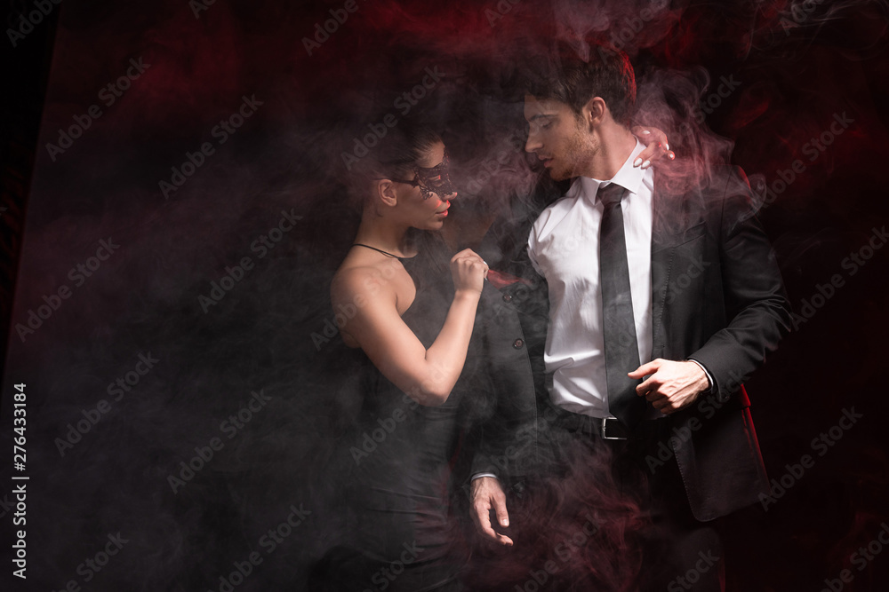 selective focus of sexy woman in mask undressing man in smoke on black