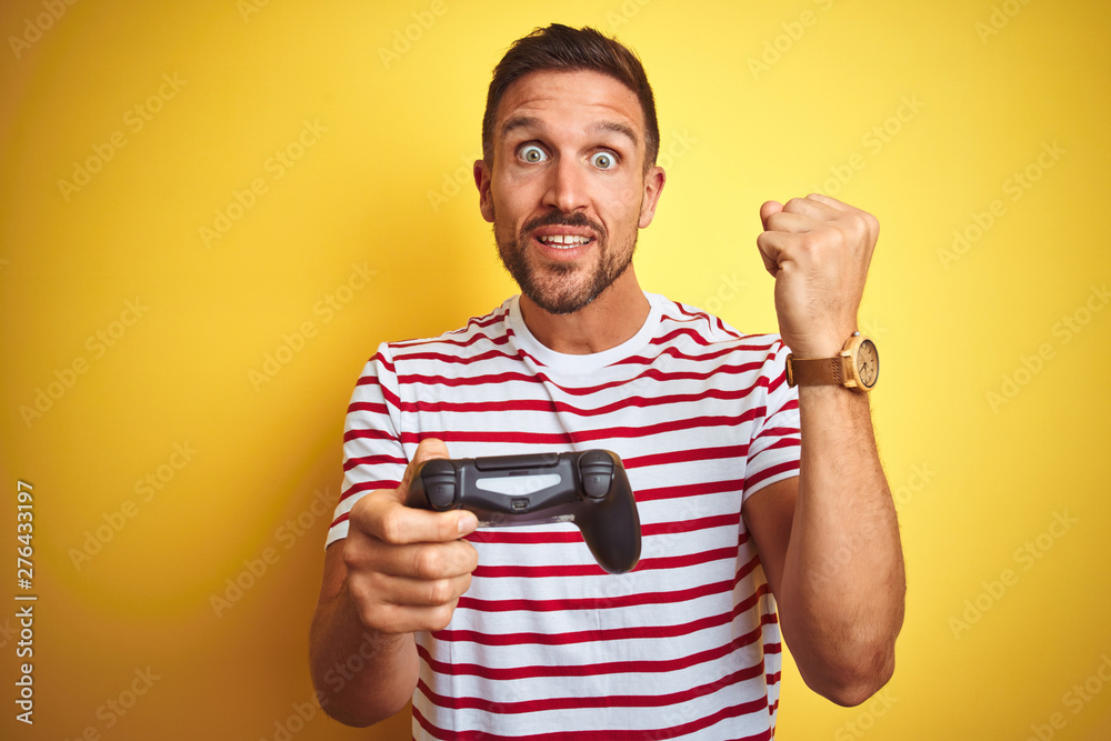Young handsome man playing video games using joystick game pad over yellow background screaming proud and celebrating victory and success very excited, cheering emotion Young handsome man playing video games using joystick game pad over yellow background screaming proud and celebrating victory and success very excited, cheering emotion