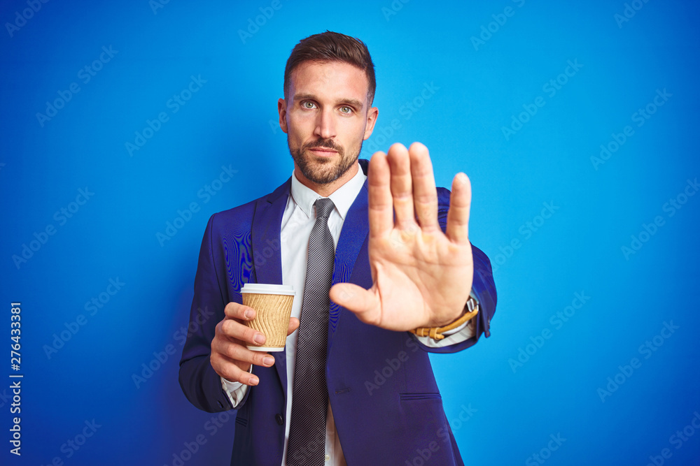 Young handsome business man drinking a coffee on a paper cup over blue isolated background with open hand doing stop sign with serious and confident expression, defense gesture Young handsome business man drinking a coffee on a paper cup over blue isolated background with open hand doing stop sign with serious and confident expression, defense gesture