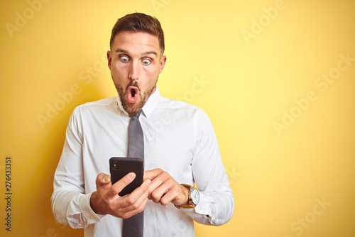 Young handsome business man using smartphone over yellow isolated background scared in shock with a surprise face, afraid and excited with fear expression