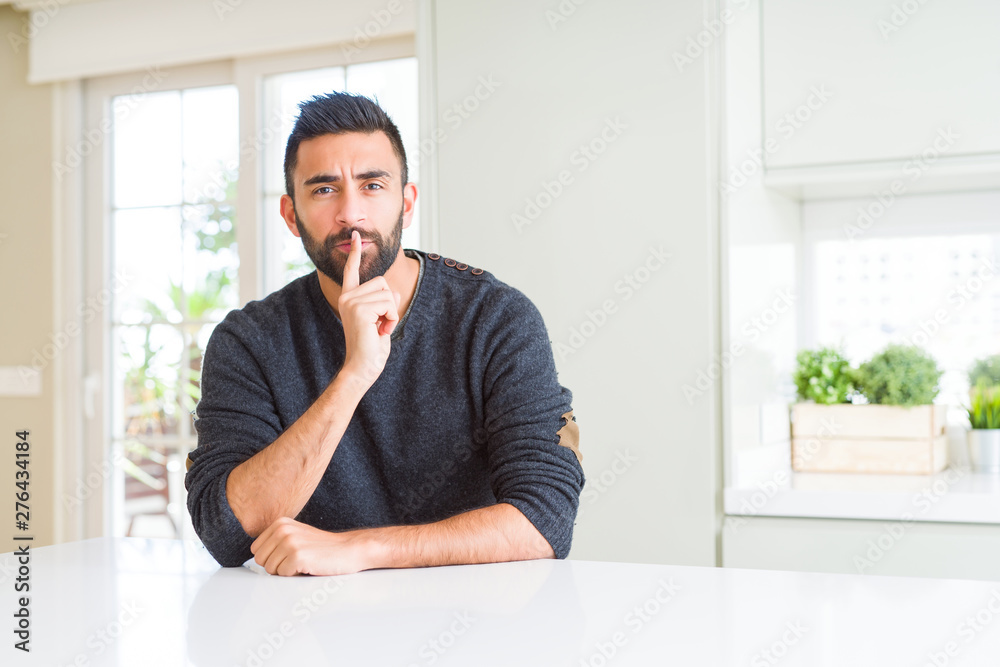 Handsome hispanic man wearing casual sweater at home asking to be quiet with finger on lips. Silence and secret concept.