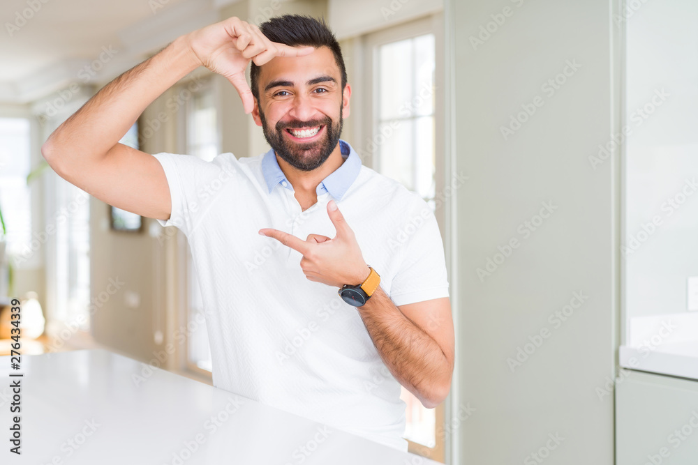 Handsome hispanic man casual white t-shirt at home smiling making frame with hands and fingers with happy face. Creativity and photography concept.